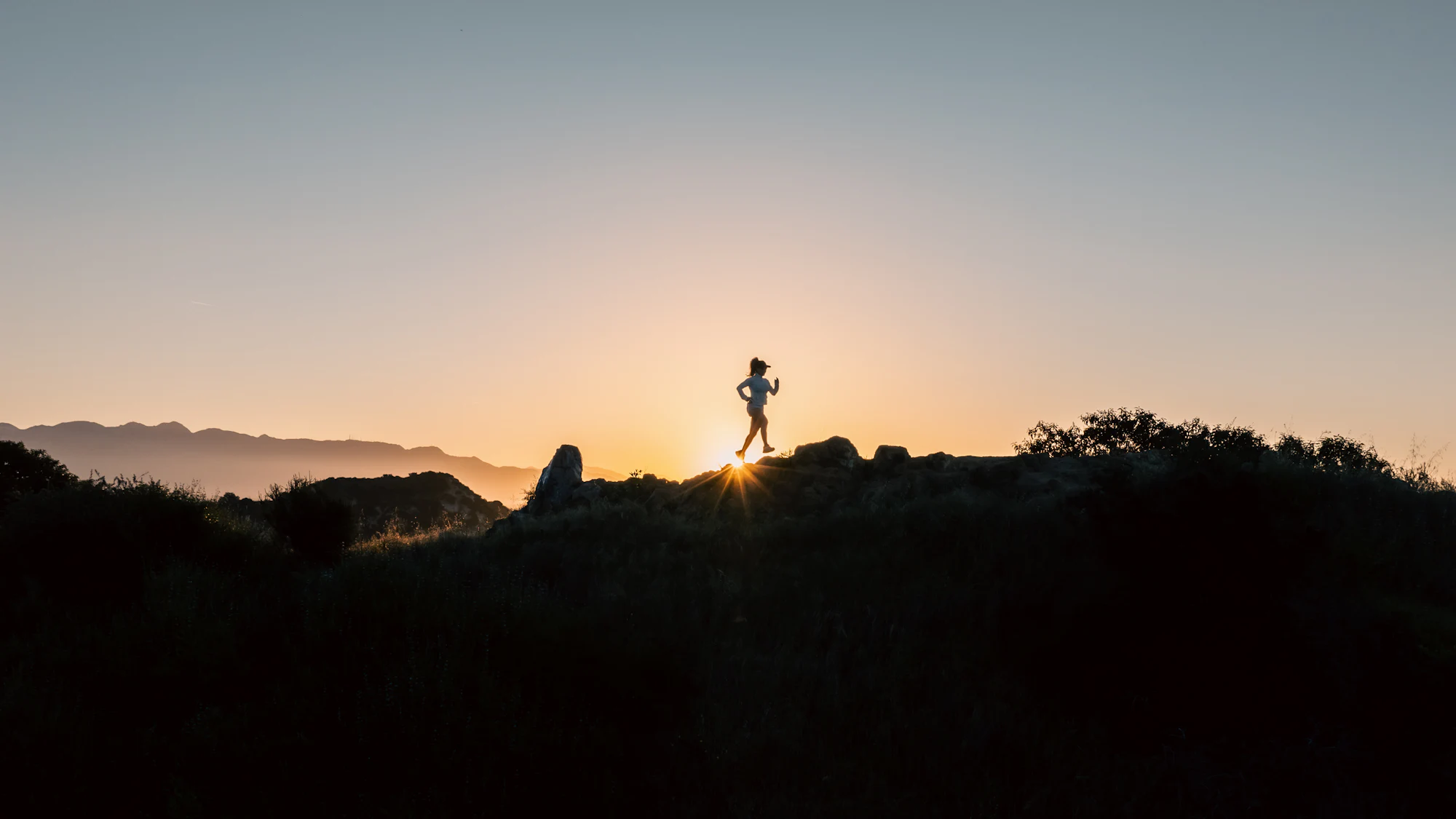 A runner crossing a rocky ridge in silhouette as the sun sets behind the mountains.